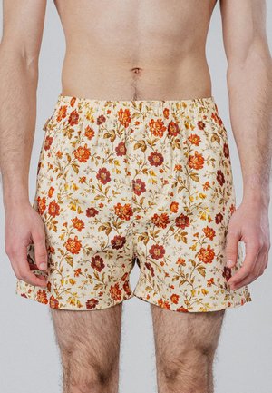 Torse et parties supérieures des jambes d’un homme portant un short couleur crème avec un motif floral orange et rouge, sur un fond clair uni.