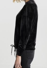 Sudadera de terciopelo negro con mangas largas, corte relajado, detalle de cordón en los laterales y un ligero brillo. Combinada con jeans oscuros ajustados.