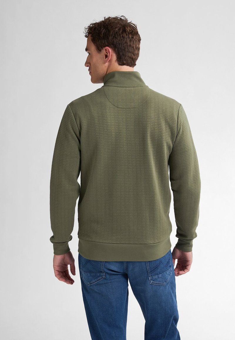 Zielona oliwkowa polarowa bluza z teksturowanym wzorem, wysokim kołnierzem i ściągaczami na rękawach, zestawiona z niebieskimi dżinsami.