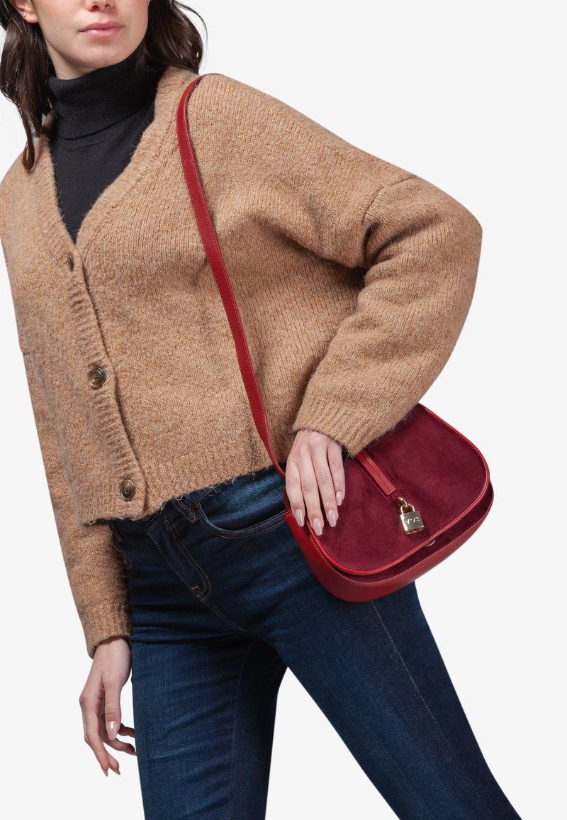 Red leather and suede shoulder bag with a curved shape, gold hardware, and a shoulder strap, worn by a model in a beige cardigan and blue jeans.