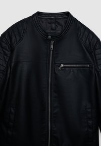 Veste en cuir noire avec un design matelassé aux épaules, fermeture éclair à l'avant et poche poitrine avec garniture métallique. Texture lisse, col rond.