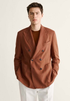 Mann trägt einen rostbraunen, doppelt geknöpften Blazer über einem passenden Hemd mit weißen Hosen, steht mit den Händen in den Taschen vor einem einfarbigen Hintergrund.