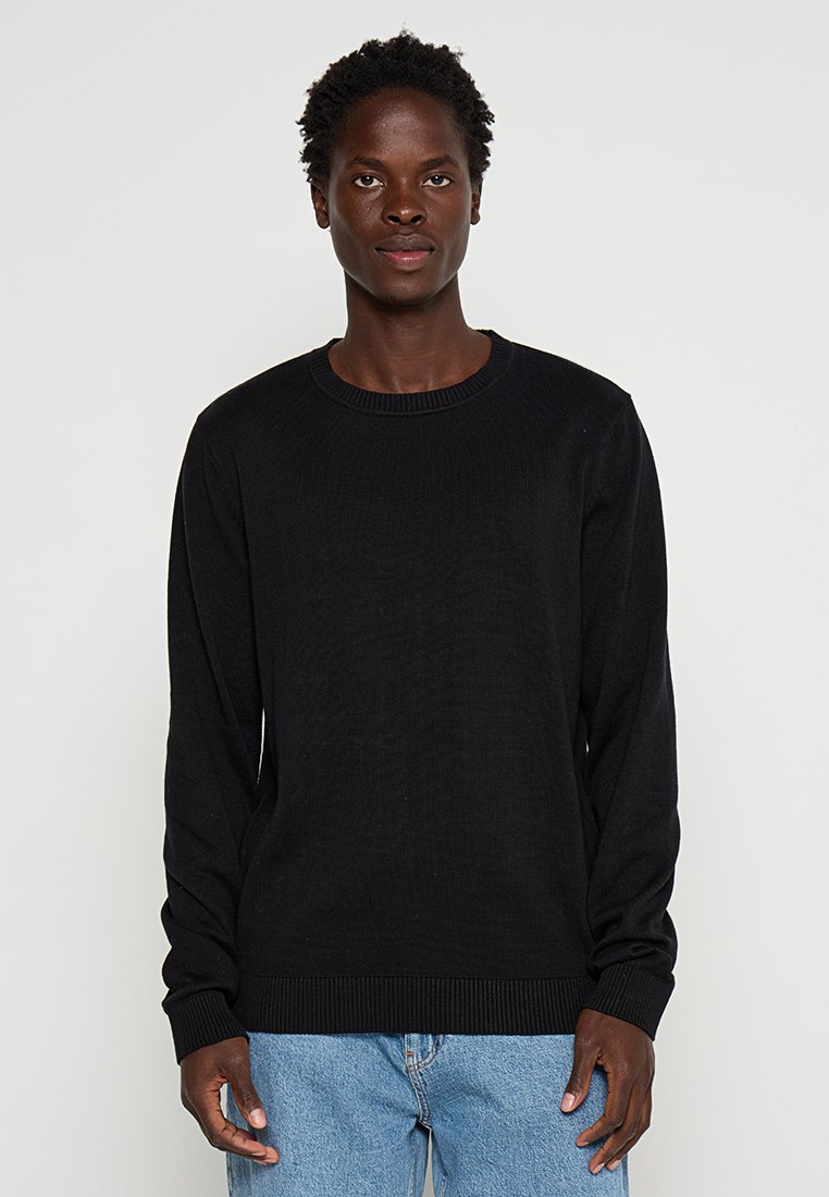 Jonge man met kort krullend haar, gekleed in een zwart crewneck trui en blauwe jeans, staande tegen een eenvoudige achtergrond.