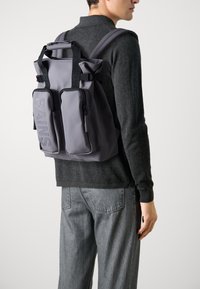 Mochila impermeable gris con múltiples compartimentos, que presenta un cierre enrollable, correas ajustables y el logo en relieve "RAINS". Textura lisa y mate.
