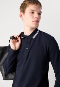 Jaunas vyras tamsiai mėlynos spalvos ilgomis rankovėmis polo marškiniais su baltais apykaklės dryžiais neša juodą odinę rankinę per petį, žiūrėdamas šoną priešais vientisą foną.