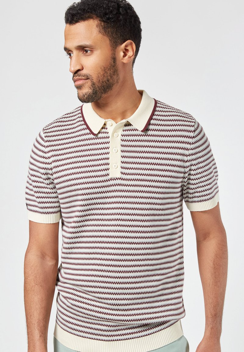 Vaaleanruskea raitapaitainen polo paita lyhyillä hihoilla, jossa on viininpunaisia ja tummansinisiä vaakaraitoja. Ominaisuuksiin kuuluu ribbauskaulus ja kolmonen napitus.