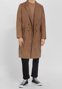 Osoba v dlouhém hnědém semišovém kabátě, hnědém tričku, černých zkrácených kalhotách a černých vysokých teniskách s bílými tkaničkami na jednobarevném pozadí.