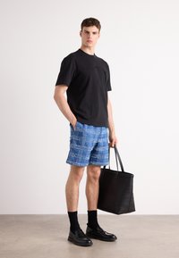 Svart kortermet skjorte med glidelås lomme, blå rutete shorts med frynser, svarte loafers, svarte sokker og en svart tote bag.