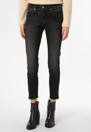 Vrouw die zwarte slim-fit jeans draagt met ruwe zoom en ritsen bij de enkels, beige geribde trui en zwarte enkellaarsjes met hak.