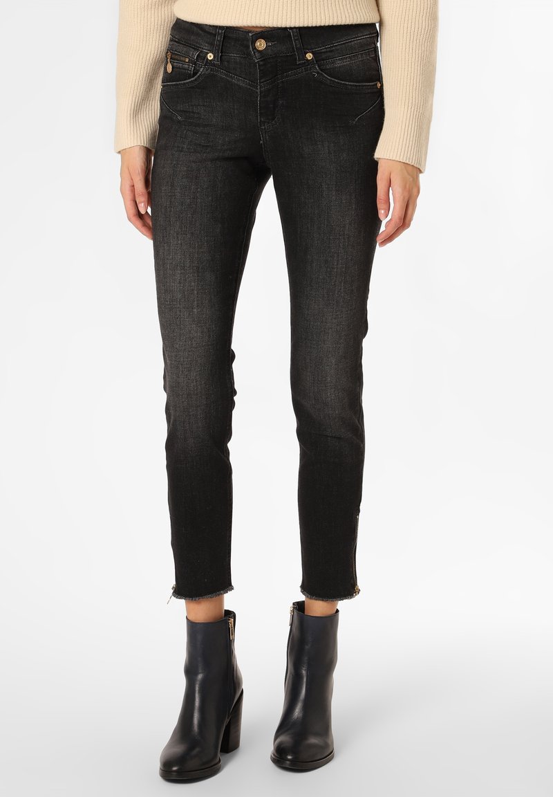 Femme portant un jean slim noir avec ourlet brut et fermetures éclair aux chevilles, un pull côtelé beige et des bottines à talons noires.
