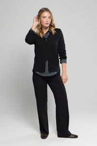 Cardigan nero sopra una camicia a righe con bottoni, abbinato a pantaloni larghi neri e scarpe nere texture. Design semplice, vestibilità su misura.