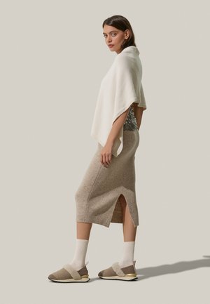 Poncho lavorato a maglia in bianco sporco, top attillato con paillettes, gonna beige con texture e spacco laterale, e scarpe slip-on beige/grigio ardesia con un accento morbido.