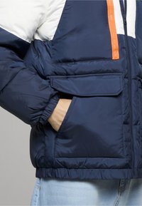 Tmavomodrá nadýchaná bunda s bielymi časťami na pleciach a na hrudi, oranžovým zipsom a prednými vreckami so zipsom na chlopni.