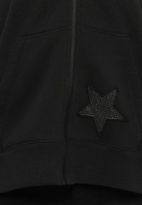 Fekete, cipzáras ruházat, elülső zsebekkel, mely a jobb zsebnél textúrált csillag alakú díszítéssel rendelkezik.