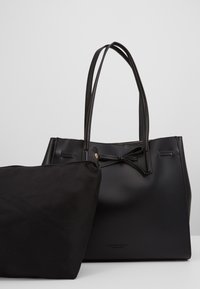 Sac fourre-tout en cuir noir avec double anses et détail de nœud, accompagné d'une pochette zippée en tissu noir sur une surface blanche.