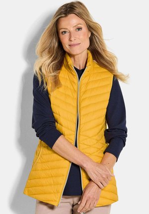 Frau mit langen blonden Haaren, die eine gelbe gesteppte Weste über einem navyblauen Langarmshirt und beigen Hosen trägt, steht vor einem weißen Hintergrund.