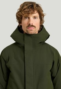 Mörkgrön vattentät jacka med hög krage och en slät textur. Har en minimalistsk design och ingen synlig hårdvara.