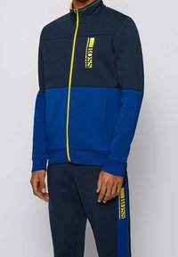 Modrá bunda na zip se žlutými detaily a logem; má vysoký límec, texturovaný materiál a kontrastní námořnické a modré části.