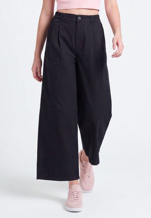 Person trägt hoch taillierte schwarze, weit geschnittene, gekürzte Hosen, ein pinkes cropped Top und hellrosa Sneakers und geht vorwärts.
