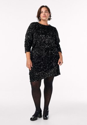 Vestido negro de lentejuelas con mangas largas, cuello redondo y cintura ajustada. Combinado con medias de lunares negras y zapatos de tacón block brillantes.