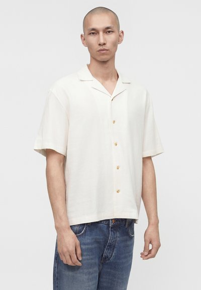 Άνδρας που φοράει κρεμ πουκάμισο με κοντά μανίκια και μπλε τζιν, στέκεται μπροστά σε απλό φωτεινό φόντο.