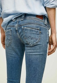 Jean en denim bleu avec une coupe ajustée, un léger délavage, des poches arrière avec des coutures incurvées, et une étiquette en cuir brun au niveau de la taille.