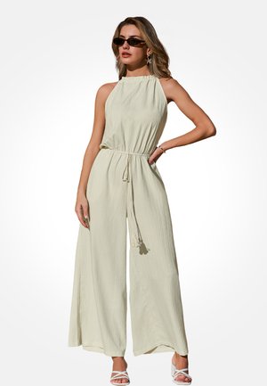 Kobieta w bezrękawnym, szerokich nogawkach kombinezonie w kolorze ecru, z talią ściągniętą sznurkiem, w okularach przeciwsłonecznych, z kolczykami w kształcie gwiazdek i białych sandałkach na obcasie.