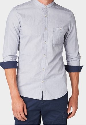 Jasnoniebieska koszula na guziki z kołnierzykiem typu mandarynka i kieszenią na piersi, rękawy podwinięte, aby odsłonić ciemnoniebieskie wewnętrzne mankiety, noszona z ciemnoniebieskimi spodniami.