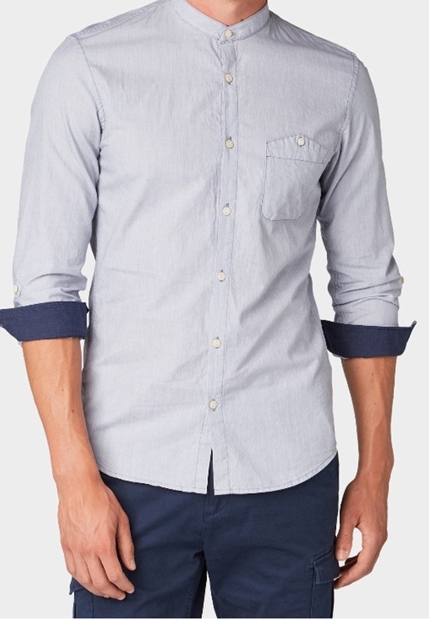 Chemise bleu clair à boutons avec col mandarin et poche poitrine, manches retroussées dévoilant des poignets intérieurs bleu foncé, portée avec un pantalon bleu foncé.