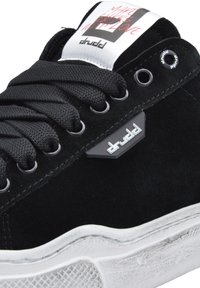 Schwarze Wildleder-Sneaker mit schwarzen Schnürsenkeln, versehen mit einer weißen strukturierten Außensohle und grauen Akzenten. Grafisches Logo auf der Zunge mit Text.