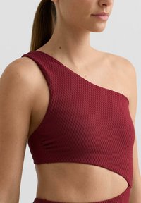 Burgundski crop top z eno ramo iz teksturiranega blaga z izrezom. Ima subtilen diamantni vzorec in prilegajočo strukturo.