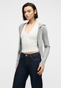 Νέα γυναίκα που φοράει ανοιχτό γκρι φούτερ με φερμουάρ πάνω από λευκό τοπ με λουράκι στον λαιμό και σκούρα μπλε τζιν, στέκεται μπροστά από απλό φόντο.