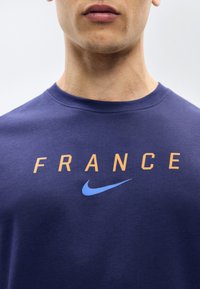 Αρσενικός κορμός που φοράει μπλε ναυτικό πουκάμισο με τη λέξη "FRANCE" σε πορτοκαλί γράμματα και ένα γαλάζιο λογότυπο Nike κάτω από το κείμενο.