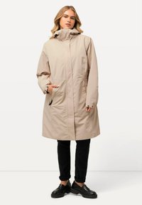 Beige wasserfester langer Mantel mit hohem Kragen, Fronttaschen und Reißverschluss. Kombiniert mit schwarzen Hosen und klobigen schwarzen Schuhen.