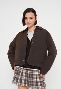 Καφέ καπιτονέ μπουφάν με κλείσιμο κουμπιών τύπου σνάπ, με δύο εμπρόσθιες τσέπες και γιακά. Φορεμένο πάνω από σκούφο πουλόβερ και καρό φούστα.