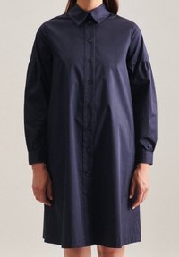 Námořnické modré košilové šaty vyrobené z bavlny. Mají límečkový krk, nabrané rukávy a mírně rozšířený lem. Jsou viditelné ploché knoflíky.