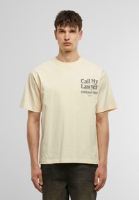 Beige bomulds t-shirt med korte ærmer og en rund halsudskæring. Har grafisk tekst på forsiden i mørkegrå. Glat tekstur.