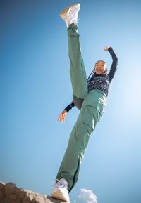 Pantalons verts légers avec poches latérales, associés à un haut à manches longues noir à motifs et des chaussures blanches. Un ciel bleu clair en arrière-plan.