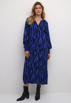 Langes blaues Kleid mit einem Wellenmuster in dunkleren Farbtönen. Verfügt über einen V-Ausschnitt, Knöpfe und geraffte Manschetten. Trägt schwarze Knöchelstiefel.