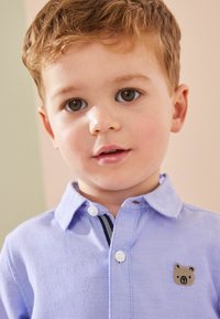 Polo shirt azzurro con colletto, caratterizzato da un logo di un orso sul petto. Il tessuto è testurizzato con una fine trama e bottoni bianchi.
