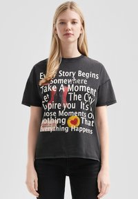 Schwarzes Baumwoll-T-Shirt mit kurzen Ärmeln, das auffälligen weißen Text und Grafiken zeigt, darunter einen gelben Kreis mit einem Herzen und einem Ticketmotiv.