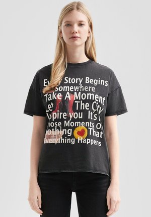 T-shirt en coton noir à manches courtes, ornée de texte et de graphiques en blanc, comprenant un cercle jaune avec un cœur et un motif de ticket.