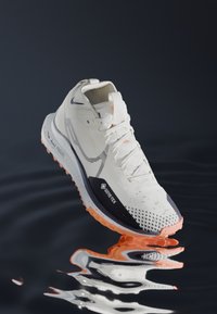 Chaussure de course GORE-TEX blanche avec accents orange, dotée d'un haut respirant, d'une semelle texturée et d'une semelle intermédiaire rembourrée pour le confort.