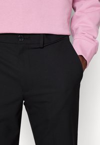 Pantaloni neri sartoriali con un design elegante, dotati di tasca laterale e chiusura con bottone. Indossati con un maglione rosa chiaro. Texture liscia.