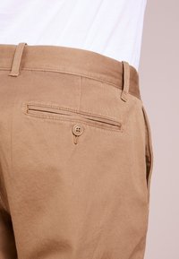 Brown cotton pants featuring a flat rear pocket with a button closure and belt loops, showing a smooth texture and tailored shape.