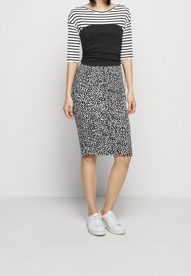 Femme portant un haut rayé noir et blanc, un milieu noir, une jupe à motifs mi-longue et des baskets blanches, debout sur un fond uni.