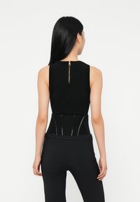 Woman with long black hair wears sleeveless black top and high-waisted black pants, standing facing away against a white background.