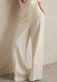 Pantaloni a gamba larga color crema realizzati in un tessuto testurizzato. Presentano una vita alta, passanti per la cintura e tasche laterali, offrendo una silhouette strutturata.