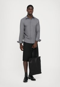 Camicia grigia a maniche lunghe con bottoni, abbinata a una gonna nera al ginocchio e a una borsa a tracolla nera. La modella è in piedi con un'espressione neutra.