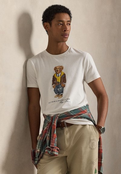 Tricou alb din bumbac cu grafică cu urs purtând o jachetă maro și o eșarfă galbenă, asortat cu pantaloni bej și o cămașă în carouri verde legată în talie.